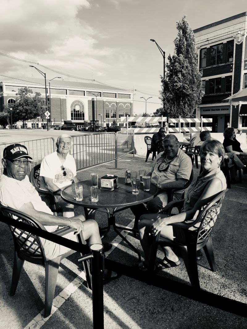 A black and white photo of four people sitting around a table on the street in Downtown Champaign. The train station is in the background. Photo by Kaitlyn Helm.