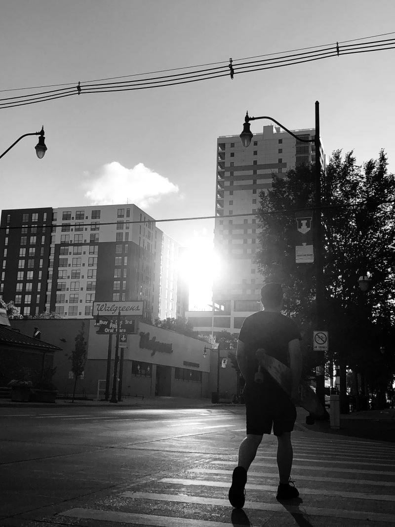 A black and white photo of a man walking along the street carrying a skateboard. The sun is shining between the high rise apartment buildings. Photo by Kaitlyn Helm.