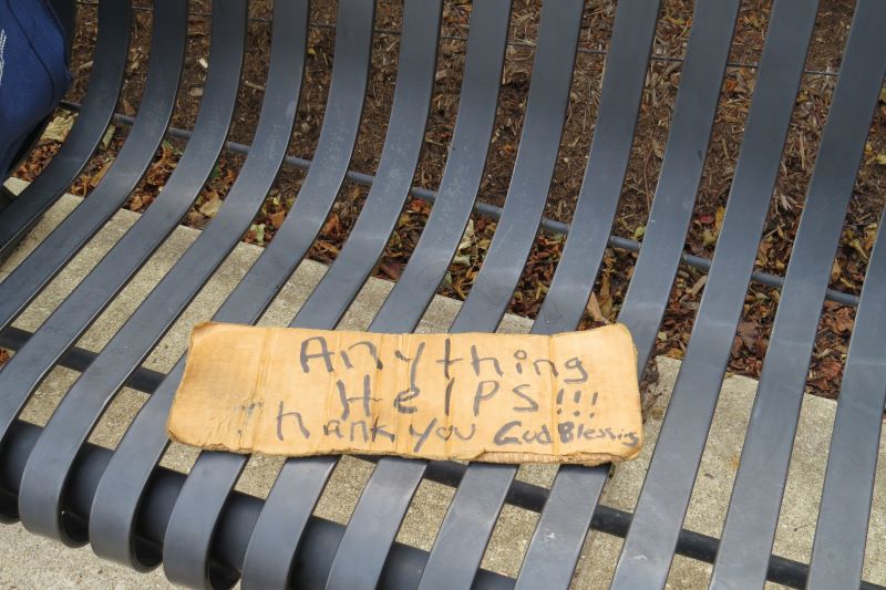 A close up of a metal park bench. A cardboard sign with the words 