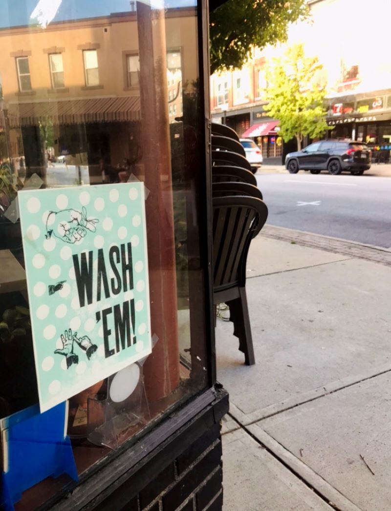 A sign that says Wash 'Em hangs in the window of a business. There is a stack of plastic chairs adjacent to the window, and a city street in the background. Photo by Grace Brock.