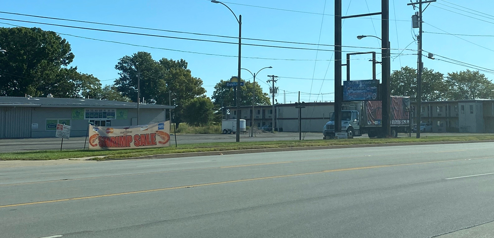 Photo of a parking lot from across a four lane road. The parking lot is empty except for a large truck. On the grass between the parking lot and the road is a sign that reads 