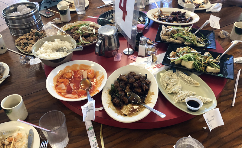 A table at Golden Harbor restaurant in Champaign is covered in many dishes of food. There are individual plates, water glasses, and tea cups, as well as many serving dishes of food on a lazy Susan. Photo by Jessica Hammie. 