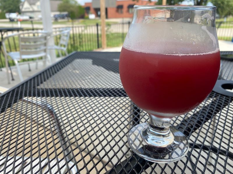 A goblet style beer glass with a reddish beer sits on a metal table outdoors. Photo by Julie McClure.