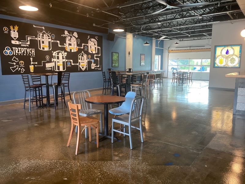 A warehouse interior with exposed ceiling and concrete floors. There are a handful of tables spread out and a garage door in the background. One wall has a painting of the beer brewing process. Photo by Julie McClure.