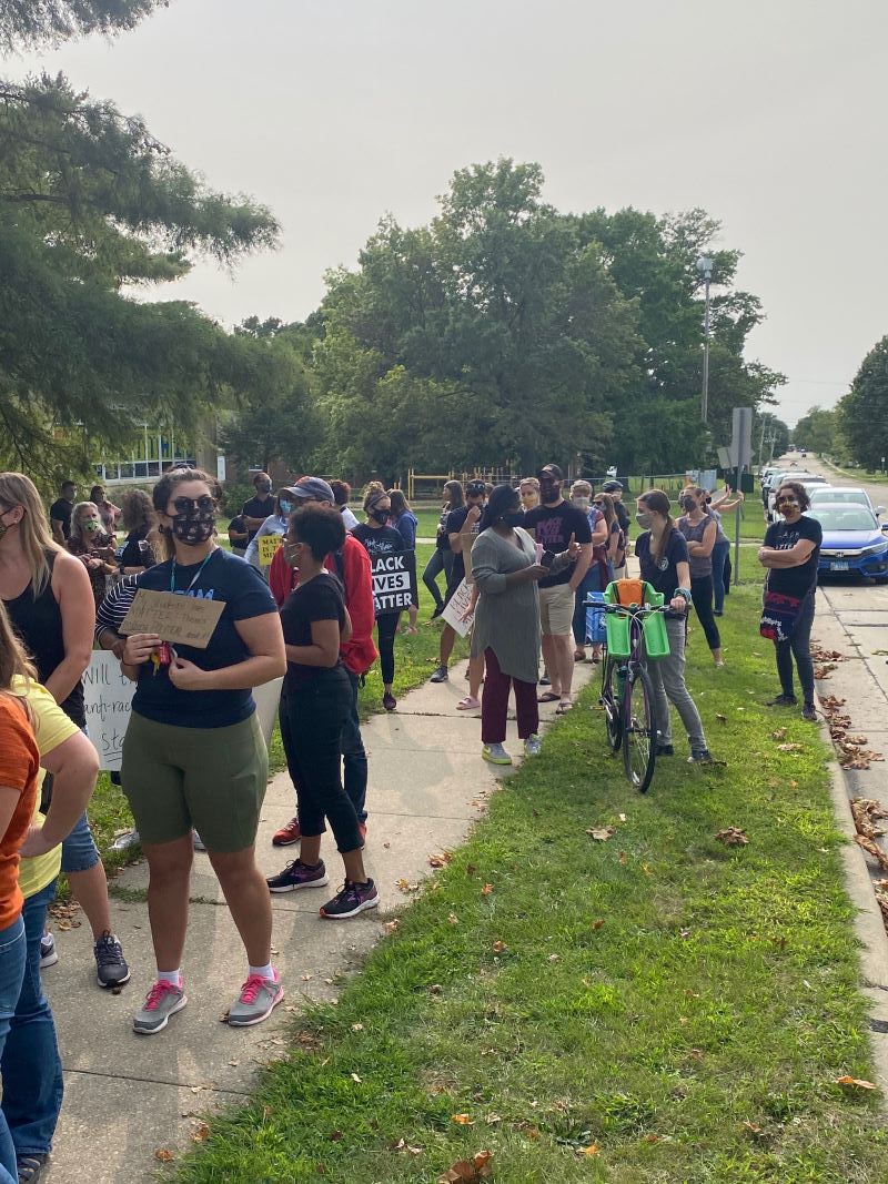 A group of people are standing along a sidewalk and in the grass alongside a school building. Some are holding signs. Photo by Julie McClure.