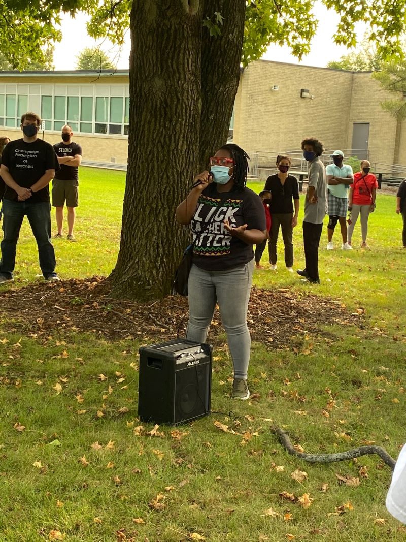 A woman wearing a shirt that says Black Teachers Matter is standing in front of a tree holding a microphone while others stand around and listen. Photo by Julie McClure.