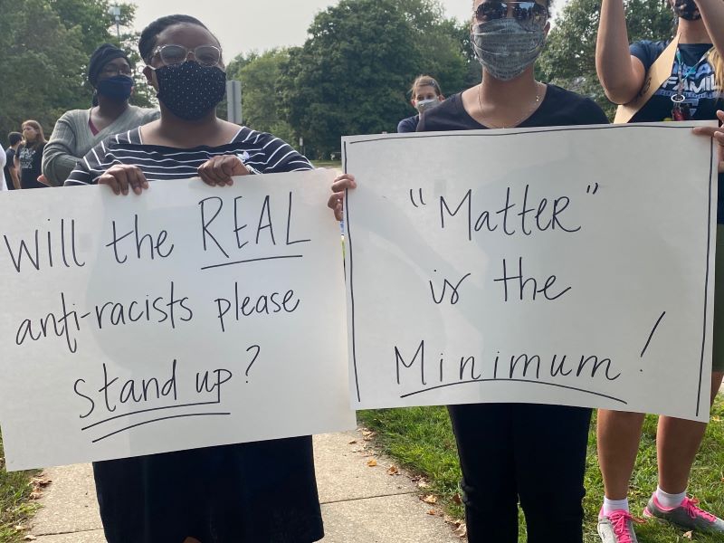 Two women wearing masks are holding white posterboard signs with black lettering. One says Will the real anti-racists please stand up? The other says Matter is the Minimum. Photo by Julie McClure.