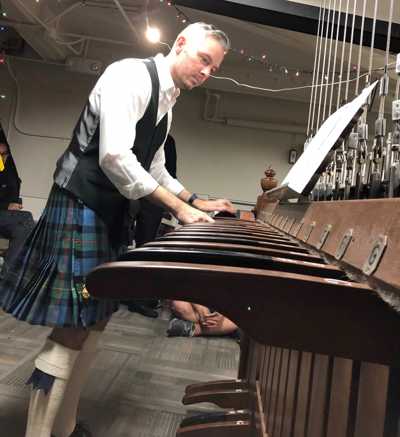 A man wearing a white collared shirt, black vest, and tartan kilt is playing the wooden keyboard that controls the chimes. Photo by Cope Cumpston.