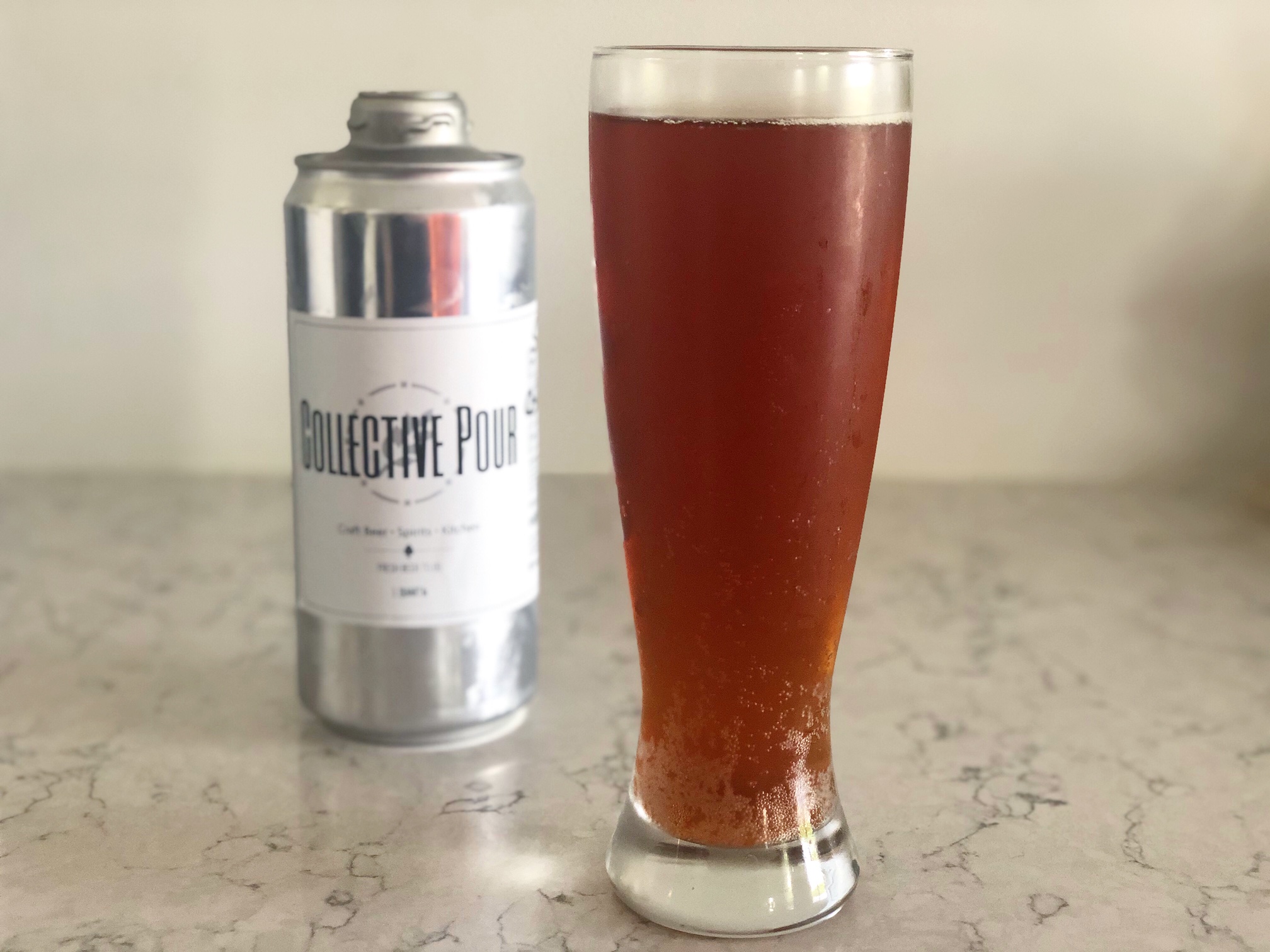 A silver crowler from Collective Pour is next to a burnt orange lager poured into a tall beer glass on a marbled counter. Photo by Alyssa Buckley.