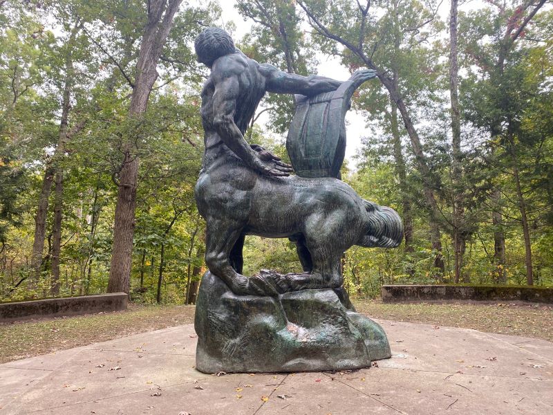 A statue of a centaur on a concrete platform. It is in a clearing surrounded by trees. Photo by Julie McClure.