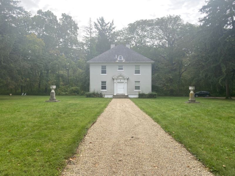 The front of a gray stucco two story house with five rectangular windows. It sits at the end of a long sidewalk. There are woods behind the house, and two stone pillars on either side of the path. Photo by Julie McClure.