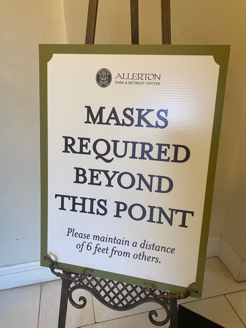 A white sign with a green border, sitting on a metal easel. It says Masks Required Beyond This Point. Photo by Julie McClure.
