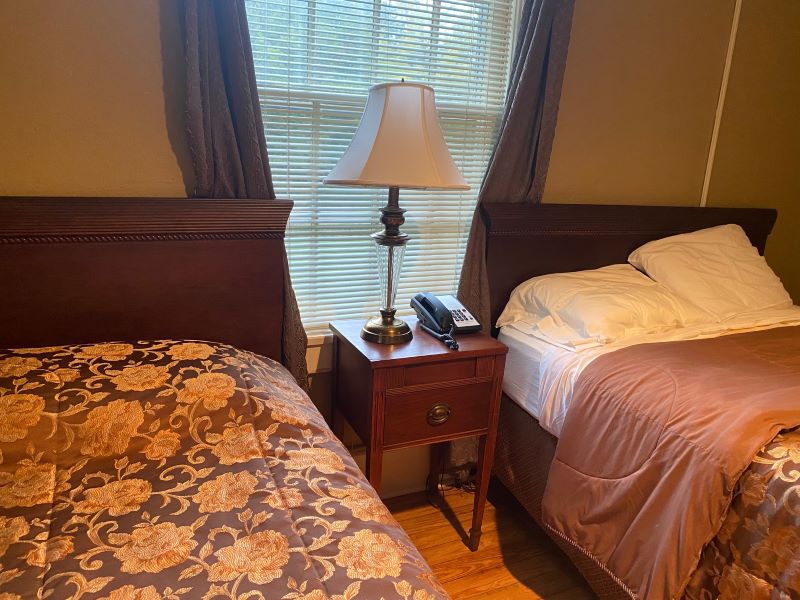 There are two beds with wooden headboards and flower patterned bedspreads. One bed is turned down, showing white sheets and pillows. Between the beds there is a nightstand with a lamp and a phone, and a window with white blinds and grayish blue curtains. Photo by Julie McClure.