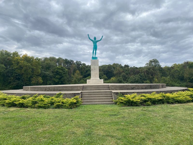 A statue of a man with arms raised. It is on a white pedestal, in the center of a concrete platform. The platform is surrounded by light green bushes, and it sits in the middle of a clearing. Photo by Julie McClure.