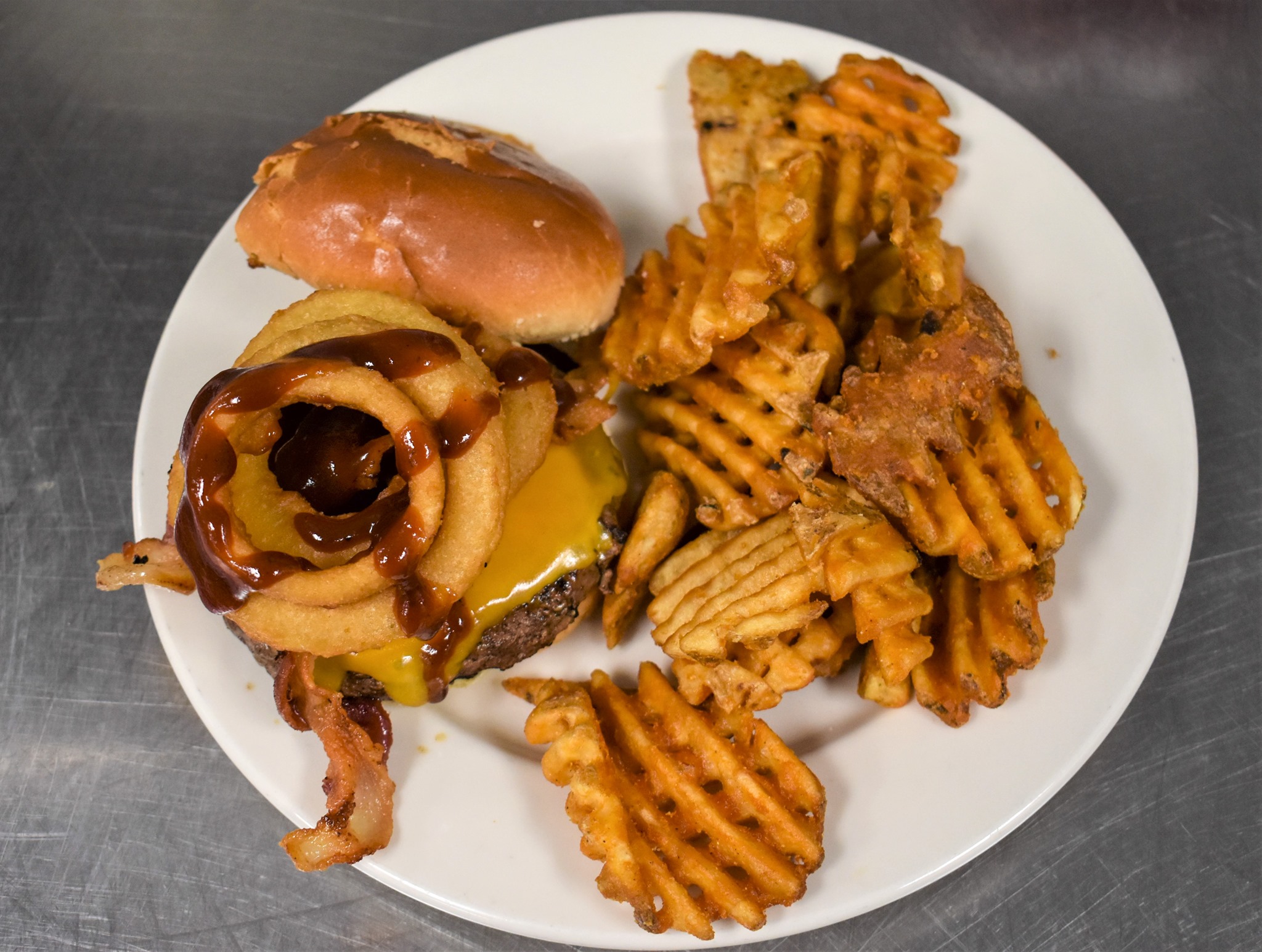 A burger with onion rings and BBQ sauce sits on a white plate with waffle fries. Photo from Aspen Tap House Facebook page.