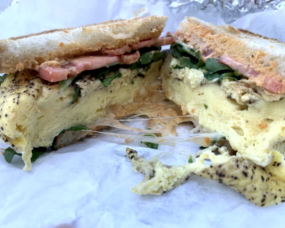 A photo of the egg sandwich from Flying Machine Avionics. The sandwich is cut in half, showing melted cheese topped with fluffy scrambled eggs, arugula, tomato slices and cream cheese. The sandwich is served on sourdough bread. Photo by Megan Friend.