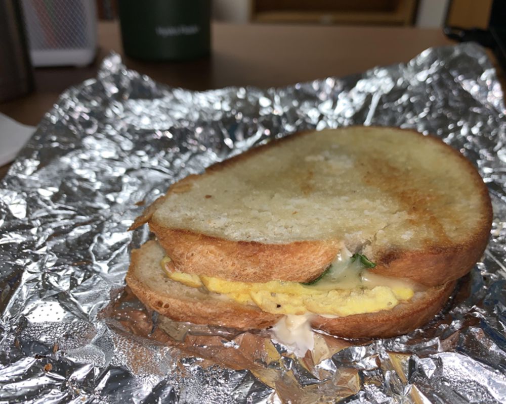 A photo of the spinach, egg and cheese sandwich from The Bread Company. The sandwich is served on buttered, toasted white bread and a small bit of spinach peaks out from inside the sandwich. The cheese is melted and on top of scrambled eggs, all sitting on a foil wrapper. Photo by Megan Friend.