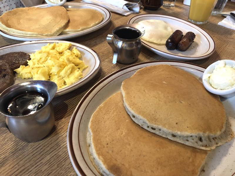 An array of breakfast dishes on white plates on a wooden table at Courier Cafe. Pancakes on two plates, eggs and sausage on the other two. Photo by Jessica Hammie.