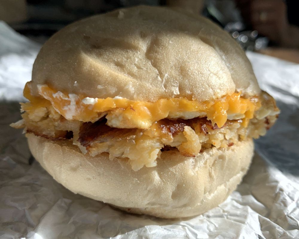 A sun-soaked photo of the Morning Bender sandwich from Cracked on Green shows a light colored french roll with layers of cream cheese, cheddar cheese and hashbrowns sitting on top of it's wrapper. Photo by Megan Friend. 
