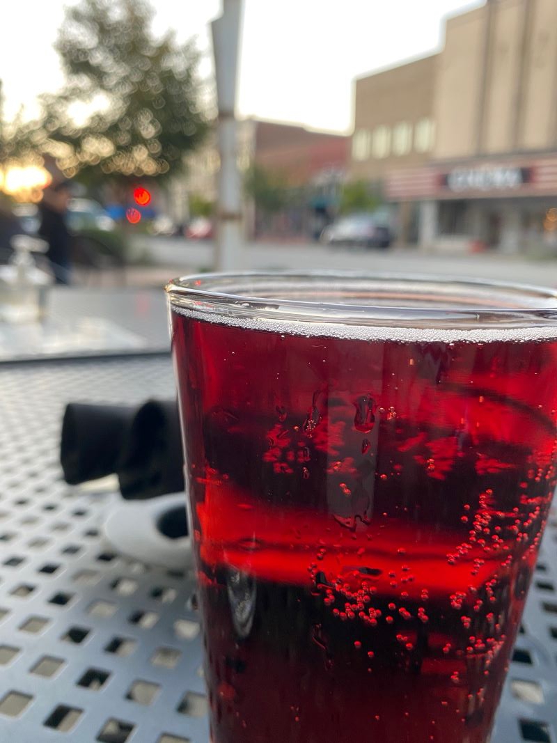 A close up of a pint glass filled with cherry red cider. It sits on a black metal table, a street and line of businesses across the street are in the background. Photo by Julie McClure.