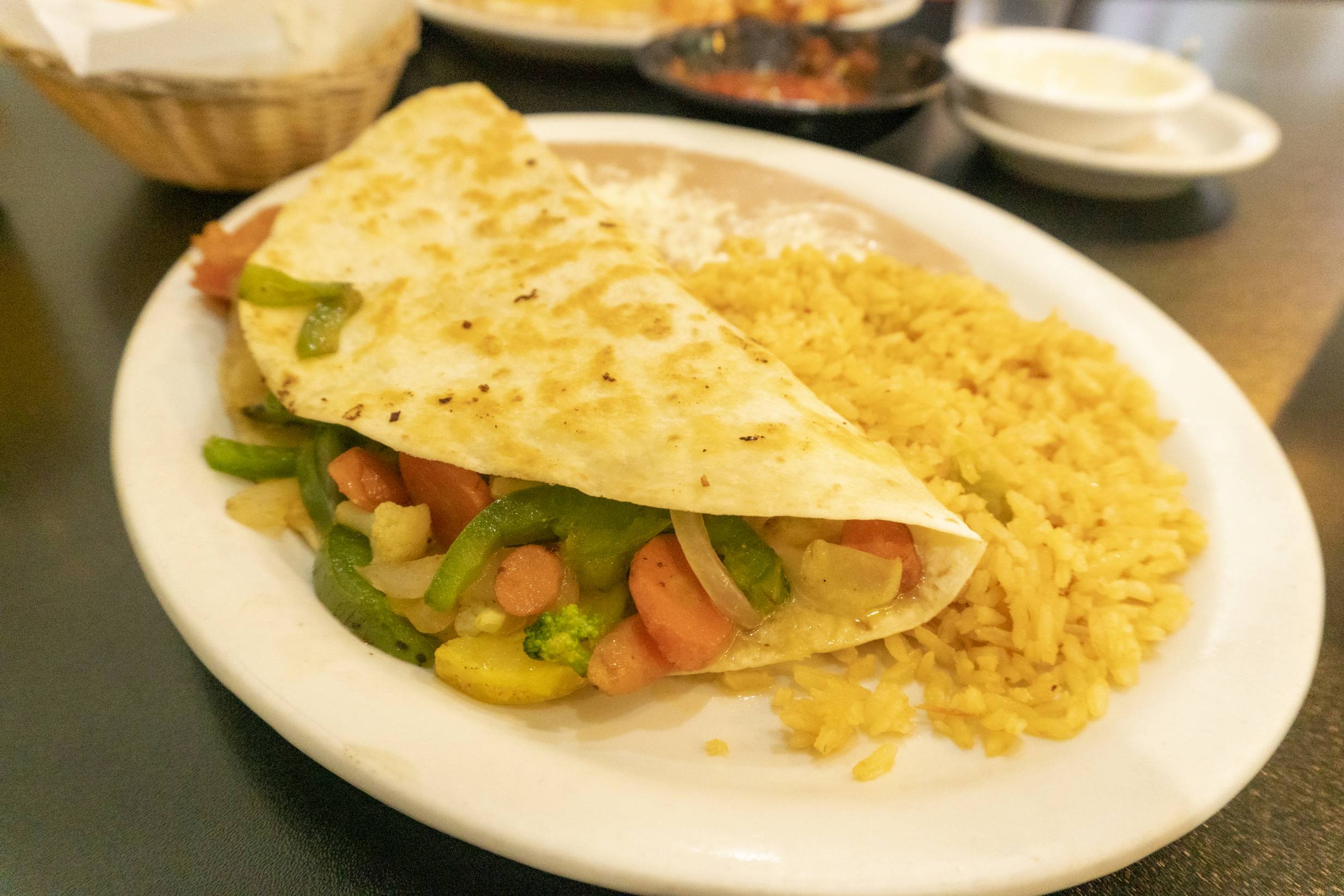 Veggie fajita quesadilla from El Patio. A crispy flour tortilla wrapped around a bed of vegatables including peppers, onions, squash, and carrots resting on refried beans and rice. Served on a white plate. Photo by Adam Rahn. 