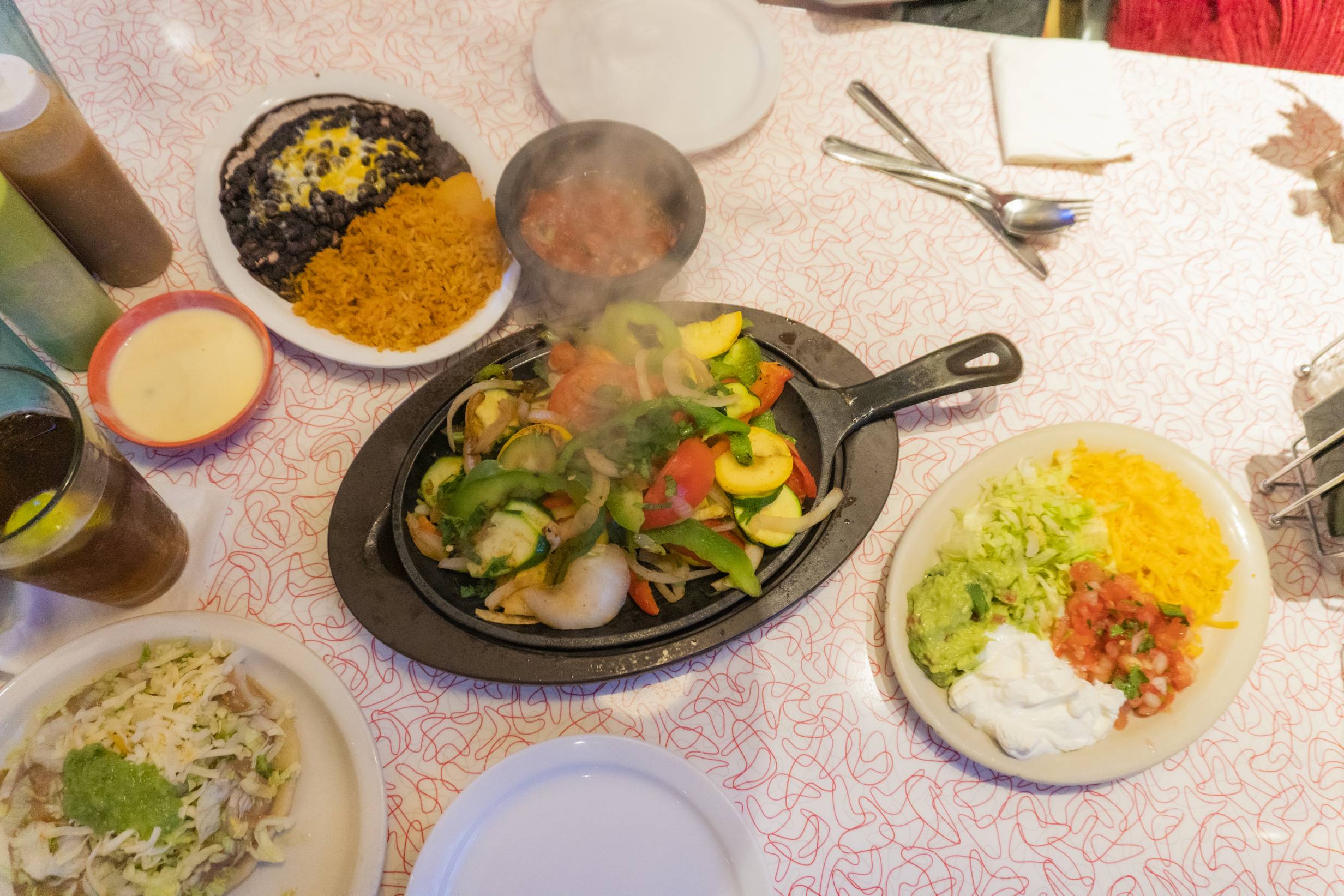 Vegetarian fajitas by Fiesta Cafe. A cast iron skillet sizzling with vegetables sits in the center. Black beans, rice, queso dip, salsa, and a plate filled with shredded cheese, lettuce, sour cream, pico de gallo, and guacamole are on the side. Photo by Adam Rahn. 