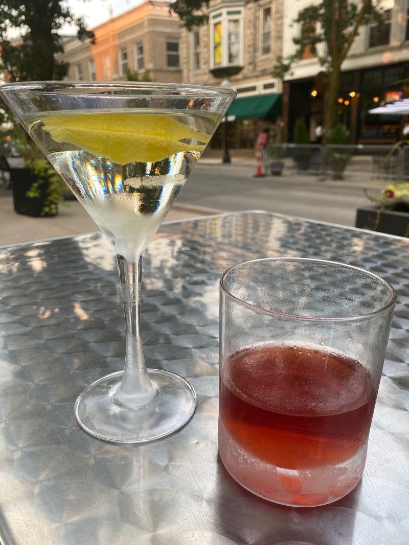 Two cocktails sit on a silver table. One is in a martini glass with a thin stem, filled with clear liquid and a lemon peel. The other is in a rocks glass with brown liquid. A street and row of businesses are in the background. Photo by Julie McClure.