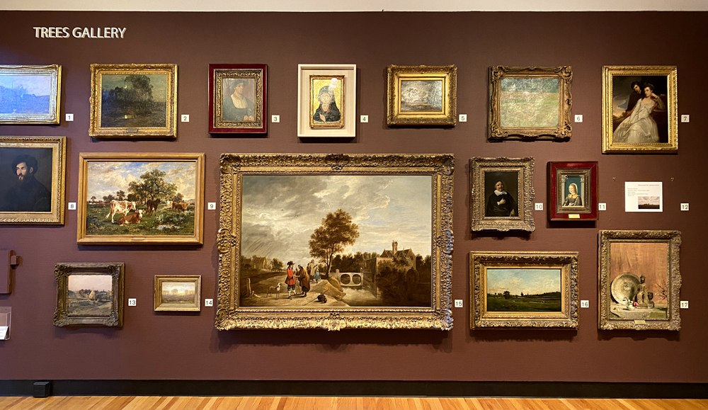 Trees Gallery in the Krannert Art Museum. The wall is painted a brownish burgundy and approximately 17 paintings are installed on one wall. Photo by Jessica Hammie. 