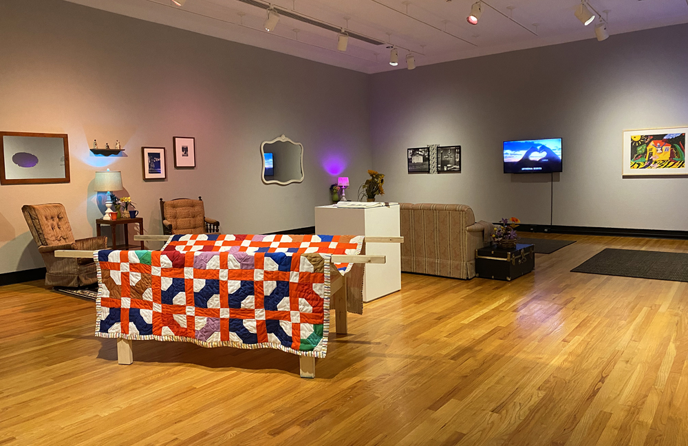 Installation view of Homemade, with Love at the Krannert Art Musuem. The installation looks like a living room, with recliner chairs, a couch in front of a wall-mounted television and various different artworks and mirrors on the wall. There is a quilt draped over a table.Photo by Jessica Hammie. 