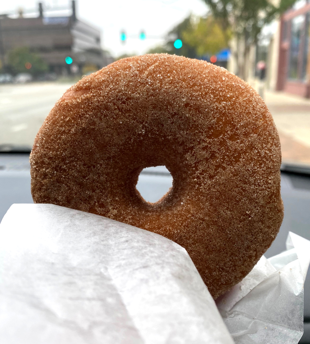 A cinnamon sugar coated yeast doughnut is held by white tissue paper. The photographer is sitting in a car, and part of the dashboard and the street are visible beyond the doughnut.. Photo by Jessica Hammie. 