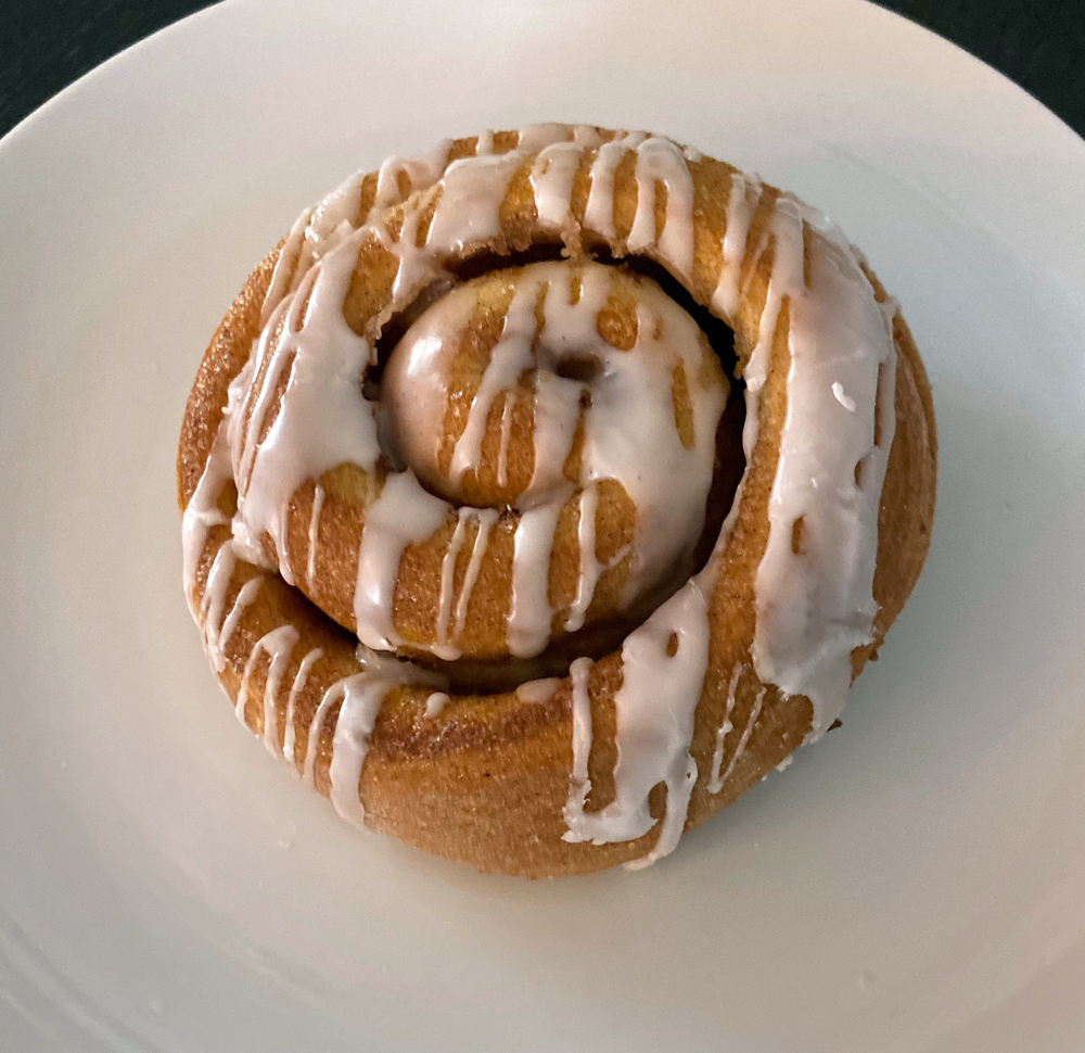 A cinnamon roll with a light drizzle of glaze sits on a white plate. Photo by Jessica Hammie. 