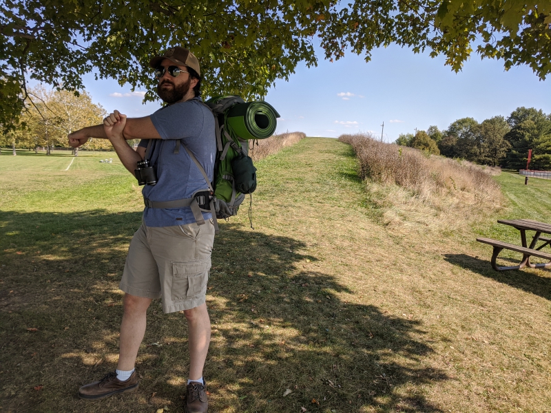 The writer is standing in front of a modest grassy hill. He is wearing a baseball cap, t-shirt, and shorts, and has a camping pack on his back. He is stretching his arm across his body. Photo by Andrea Black.