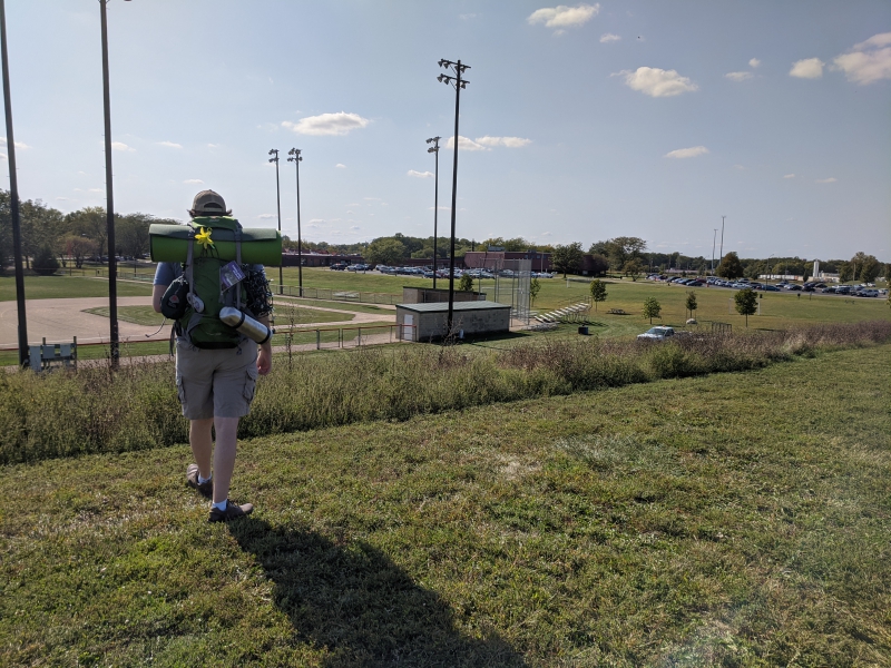 The writer is standing with his back to the camera, overlooking a baseball field and parking lot full of cars. Photo by Andrea Black.