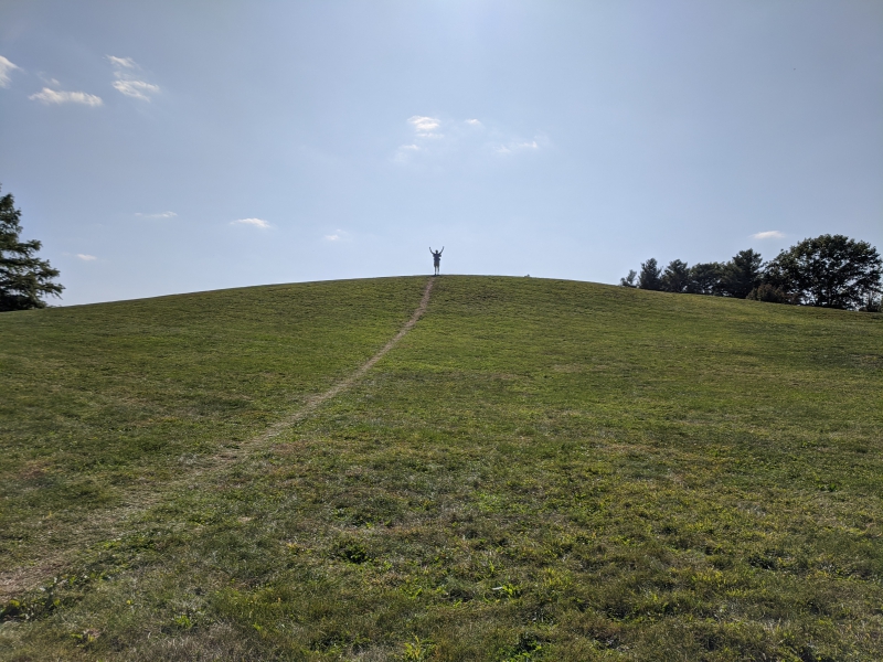 The writer is way off in the distance at the top of a grassy hill, raising both arms in the air. There is a thin dirt path that runs up the length of the hill. Photo by Andrea Black.