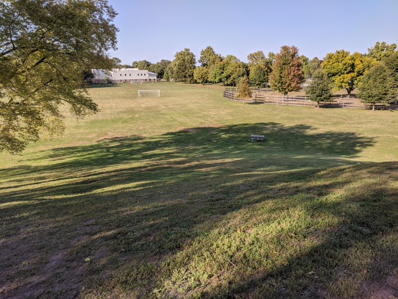 A view down a grassy hill. A white soccer goal is at the bottom of the hill, and there is a white and light brown rectangular building in the background. Photo by Andrea Black.