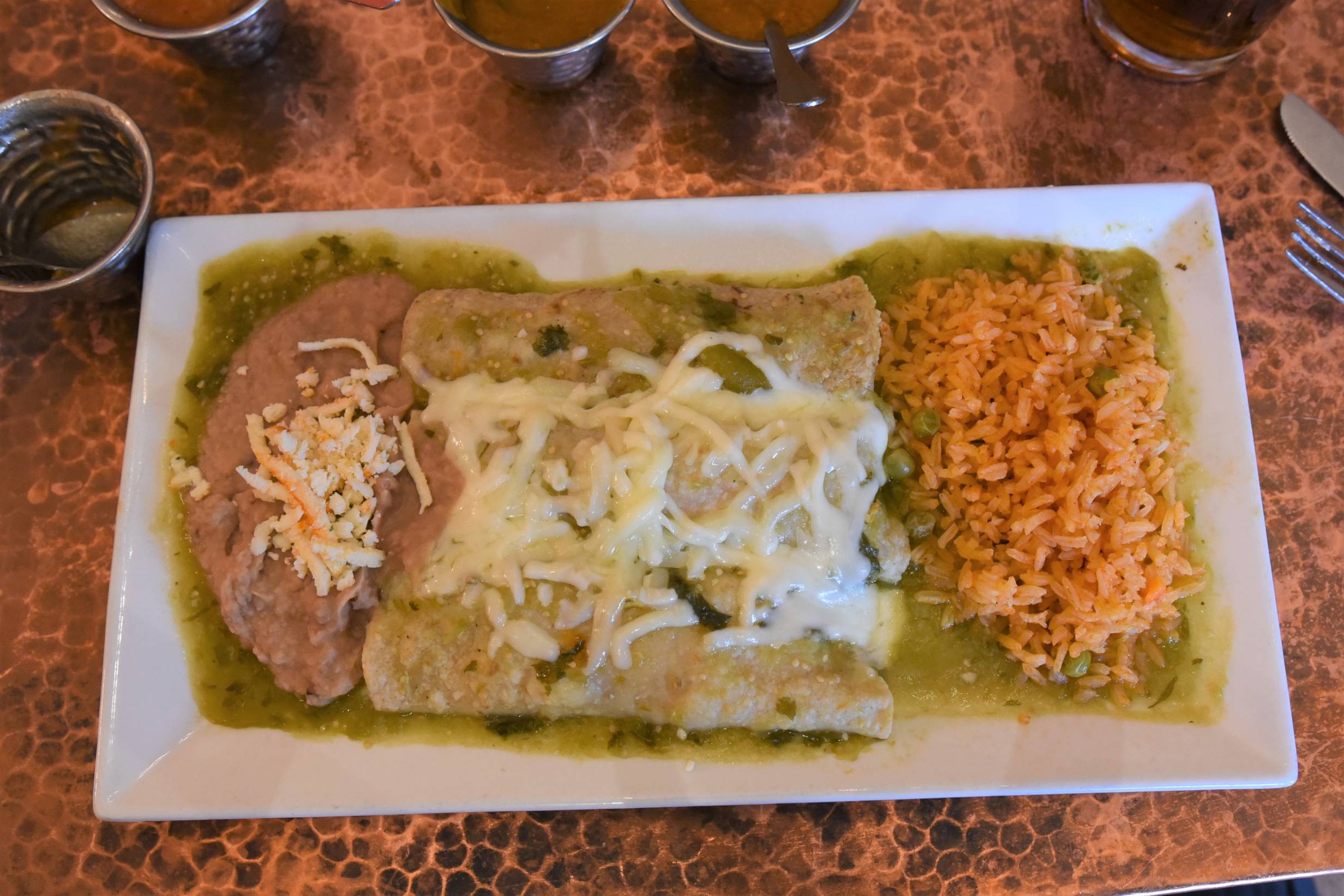 Enchiladas en Salsa Verde from Maize. An overhead photo of three cheese and zucchini blossom enchiladas covered in green salsa and topped with melted white cheese. Refried beans and rice are served on the side. Served on a white plate. Photo by Jordan Goebig. 