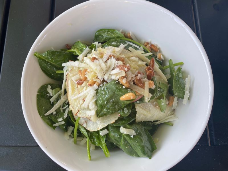 A spinach salad in a large white bowl, topped with shredded cheese, thinly sliced pears, and toasted brown pine nuts. It sits on a dark gray table. Photo by Julie McClure.
