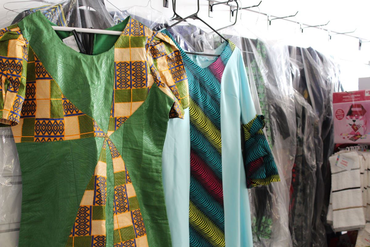 A parrot green tunic with yellow accents and a sky blue tunic with yellow, purple, and darker blue accents hang on a wall.  Other clothing in plastic wrappings can be see behind them and to their right. Photo by Tias Paul.