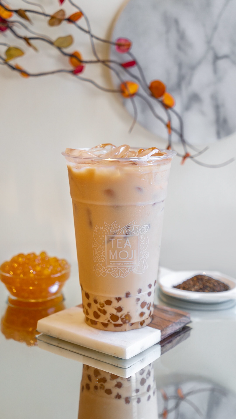 A white drink with dark boba at the bottom in a plastic cup sits on a white square coaster with fall leaves in the background. Photo by Teamoji. 