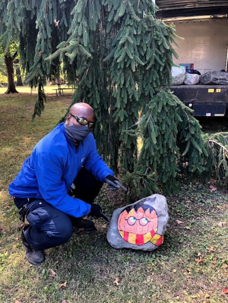 A man in sunglasses and a mask is kneeling next to an evergreen tree, pointing at a rock with a Harry Potter pumpkin painted on it. Photo from Champaign Park District Virtual Fun Facebook page.