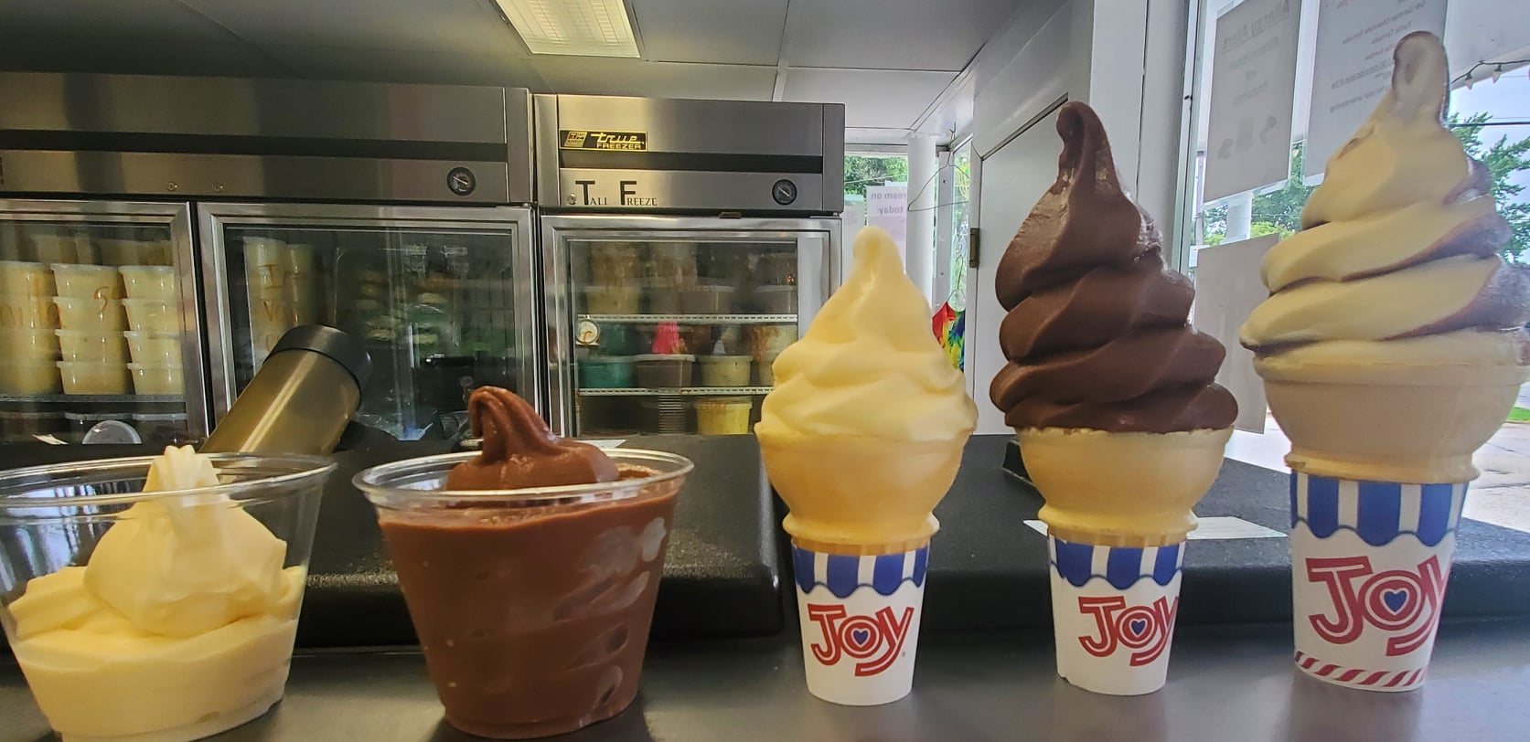 A vanilla ice cream in a cup sits next to a larger chocolate ice cream in a cup. Beside them to the right are three ice cream cones. Photo from Sidney Dairy Barn's Facebook page.