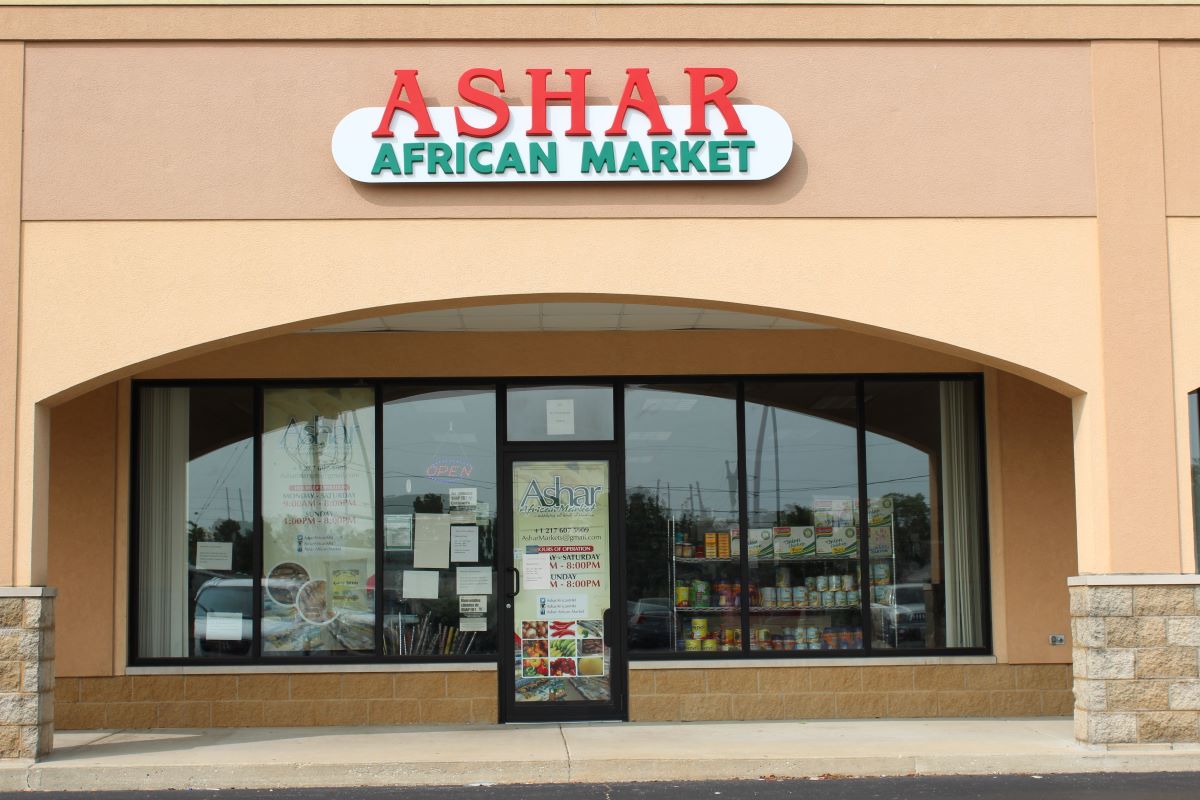 An image of the store front from the parking lot.  The glassfront doorway is framed by a flesh-colored archway, flanked by concrete and masonry pillars.  Above the archway, is the name of the store in bold lettering, Ashar in red, African market in green. Photo by Tias Paul.