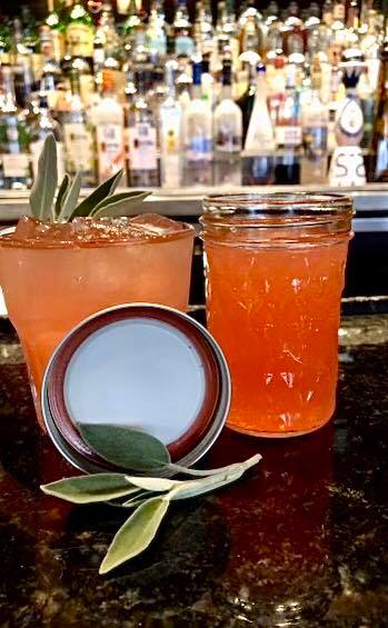 On Seven Saints' bar, there is a pink drink called Flight of Icarus in a half tumbler and also in a mason jar with the lid removed. A sprig of sage leaves sits beside the two cups. Photo from Seven Saints' Facebook page.