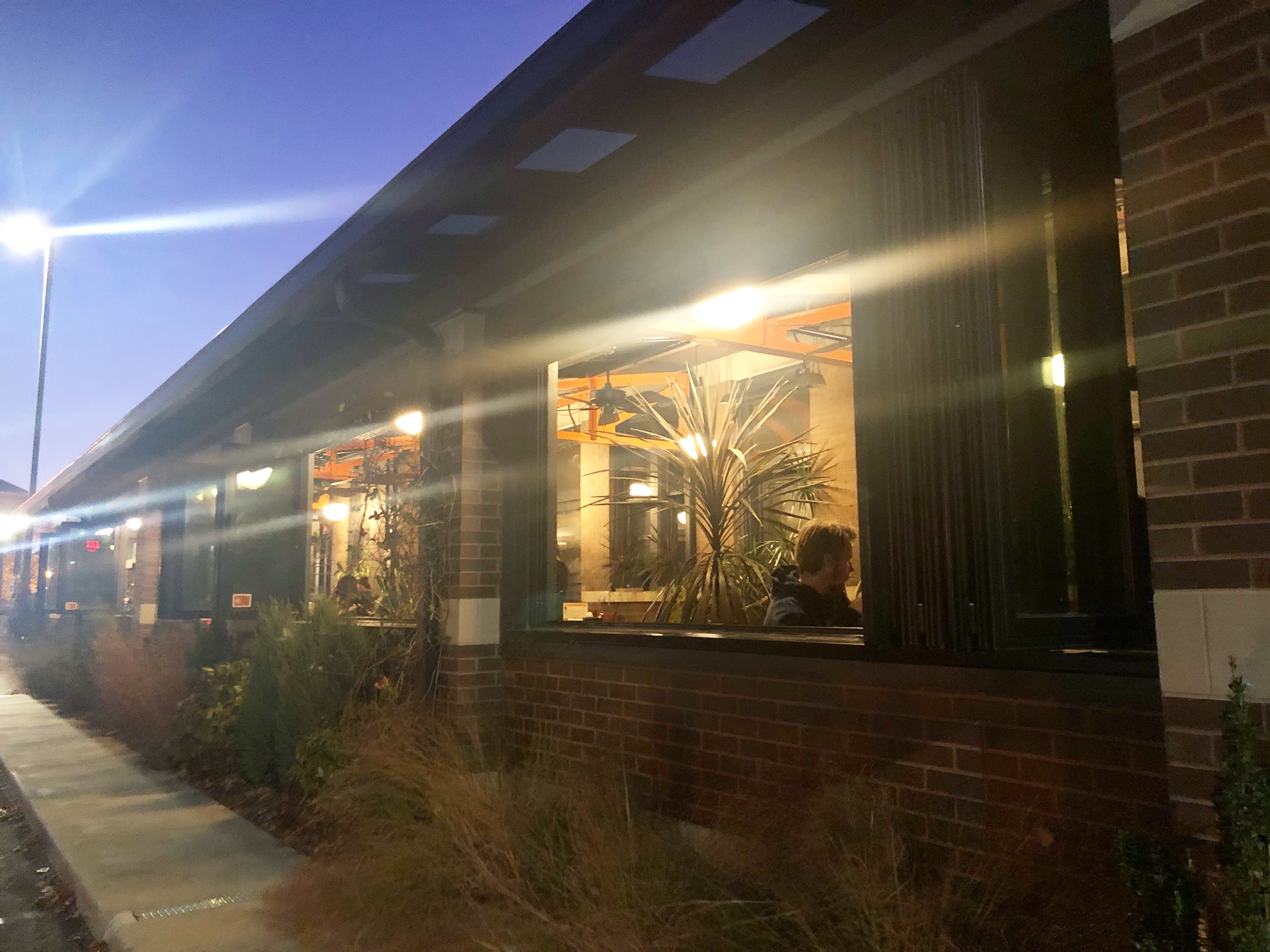 An exterior photo of Black Dog Champaign's four seasons porch. There is a male diner at one of the tables inside with the windows open on a dark night. Photo by Alyssa Buckley.