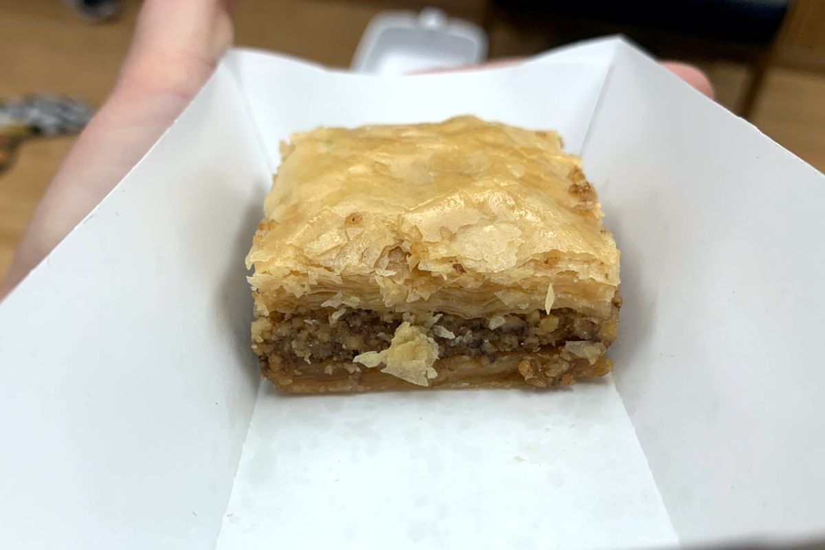 Photo of a square piece of baklava. There are layers of filo dough on top of a layer of finely chopped walnuts and layers of filo dough on the bottom. The baklava is in a paper food tray. Photo by Megan Friend.