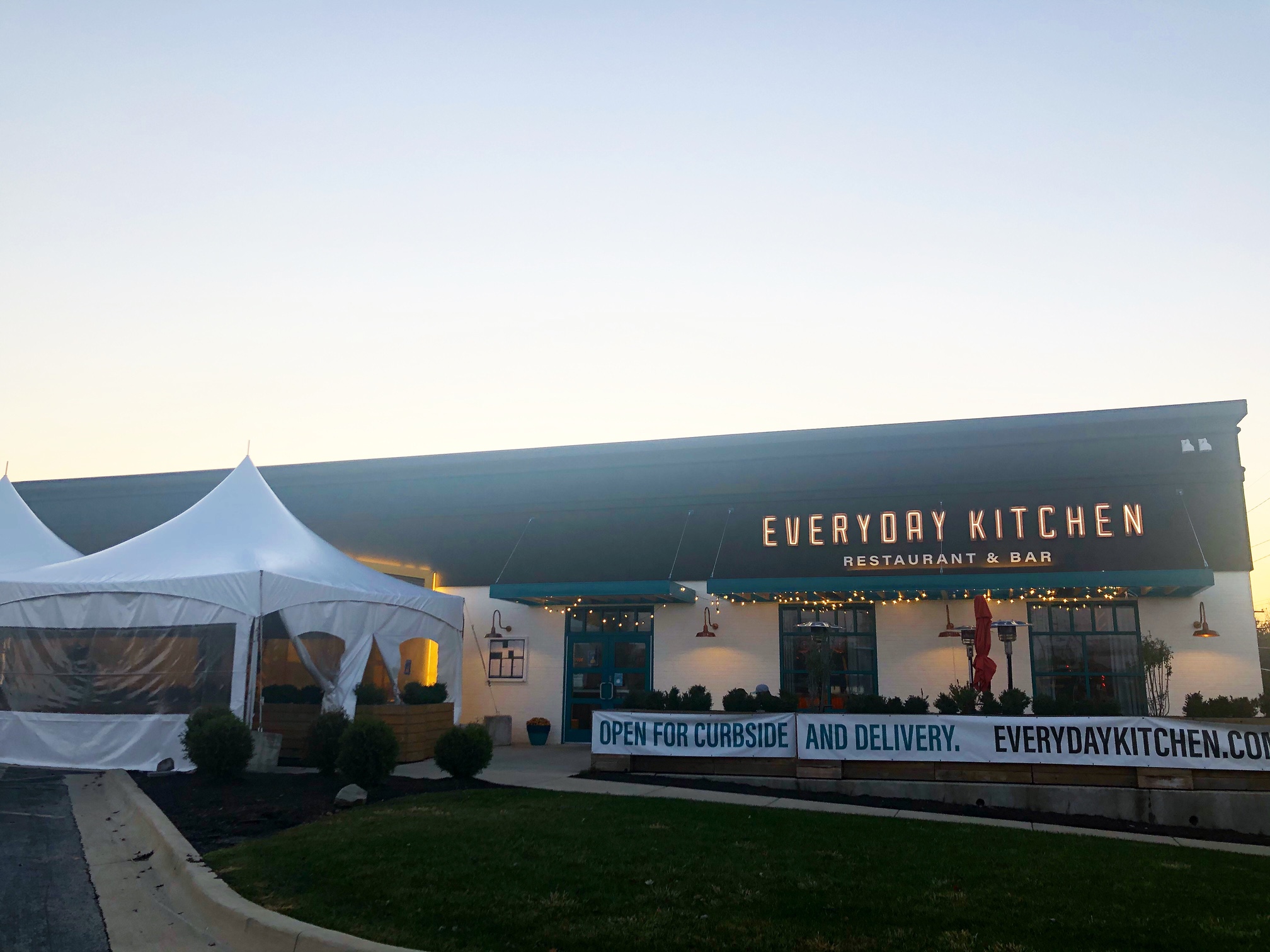 The exterior of Lodgic Community in Champaign's Everyday Kitchen Restaurant is lit up from the inside at sunset. Outside, there is a giant white tent with panoramic windows on the left and tables spaced apart on the patio. Photo by Alyssa Buckley.