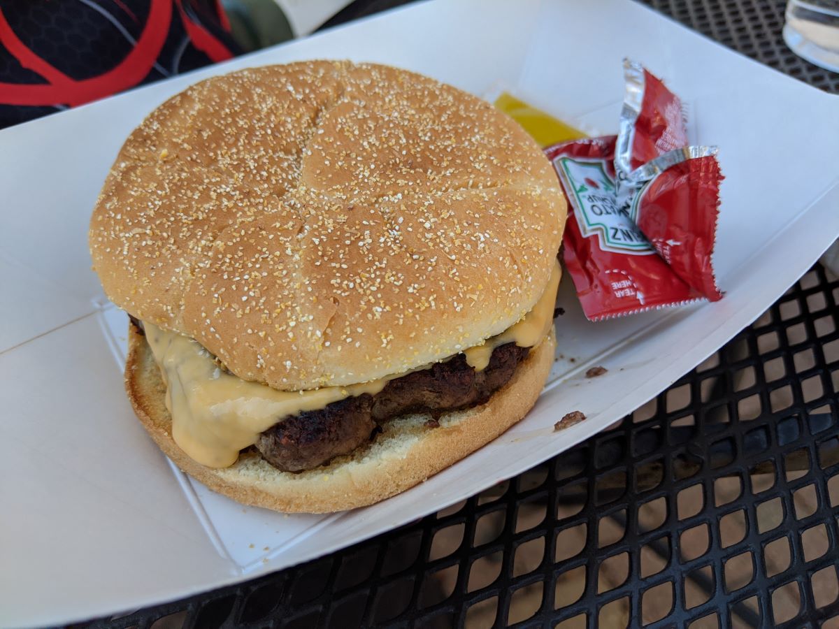 An order of the 1/3lb cheeseburger from the Huberâ€™s menu. The burger is served in a small paper tray with packets of ketchup and mustard on the side.  Below the tray, we see the metal top of a patio table. Photo by Tias Paul.