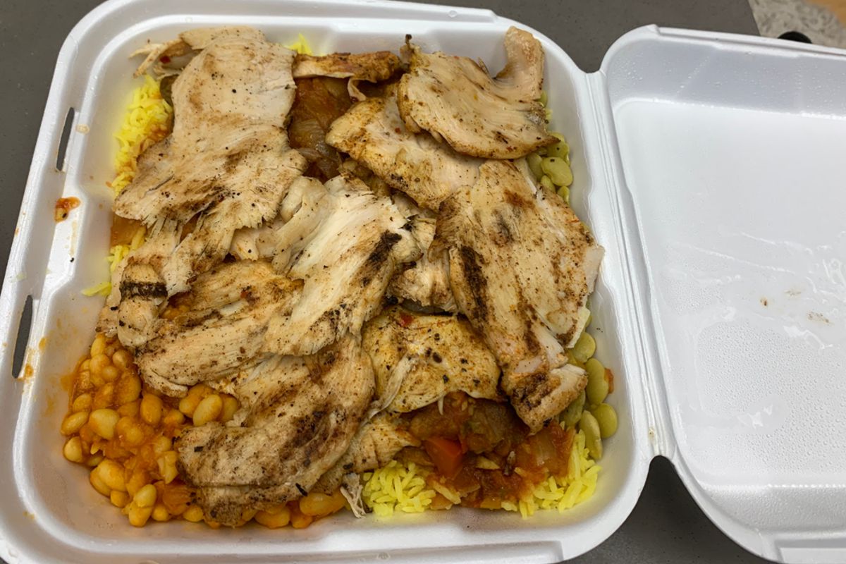 A photo taken from above of a Styrofoam takeaway container filled with yellow rice covered with thin sliced of cooked chicken. There are two types of beans underneath the chicken, a white bean in an orange sauce and green fava beans. There is a red salsa looking sauce underneath the chicken on top of the rice. Photo by Megan Friend.