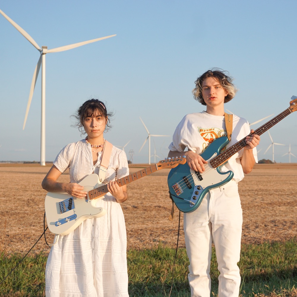 A woman (pictured left) holds an electric guitar wearing a white dress next to a taller man wearing all white, holding a bass guitar. In the background is a blue sky and wind turbines. Photo by Soft and Dumb.