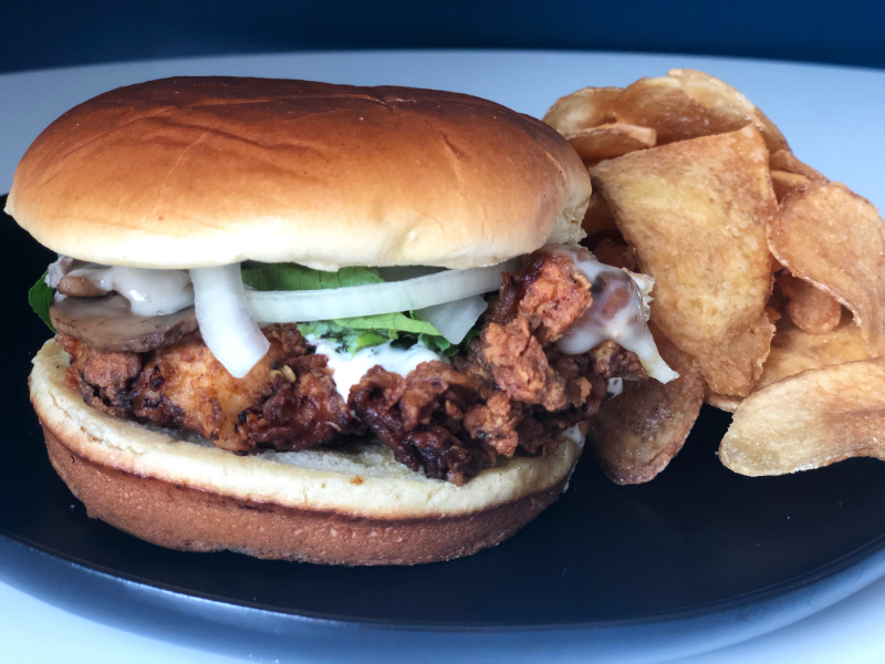 A thin fried chicken sandwich has cheese covered mushrooms with raw onion and lettuce on it on a black plate on a white table. Photo by Alyssa Buckley.
